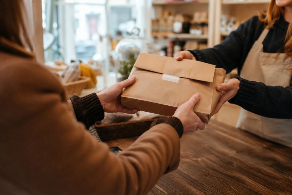 A shop worker in an apron handing a brown-paper-wrapped order across the counter to a collecting customer